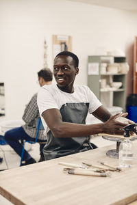 Young man wearing apron molding clay while sitting at table in classroom