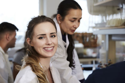 Portrait of smiling young female student sitting against teacher friends in chemistry laboratory