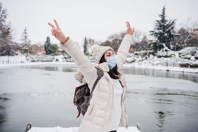 Happy backpacker woman wearing face mask standing on pier in front of frozen lake in city. 