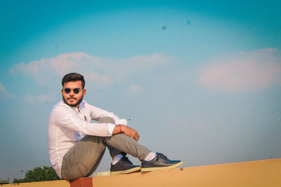 Full length of young man sitting on retaining wall against sky