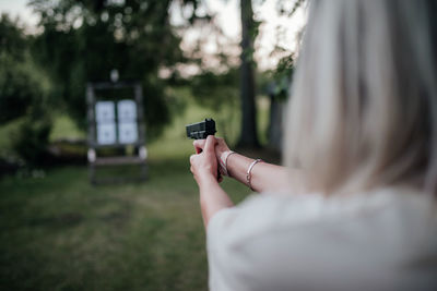 Young blonde adult woman holding gun and shooting to target in countryside at daylight. rear view