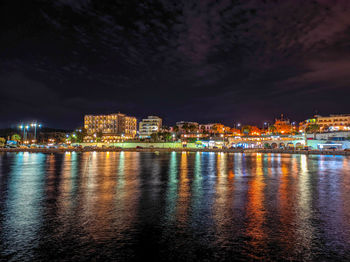 Illuminated buildings by river against sky at night