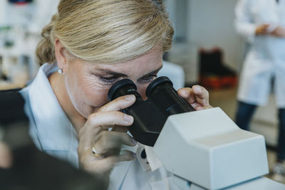 Female scientist looking through microscope while man standing in background at laboratory