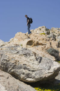 Low angle view of teenage boy standing on rocky mountain against clear blue sky