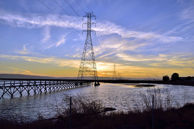 Silhouette electricity pylon by bridge against sky during sunset