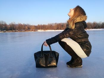 Side view of smiling woman with shoulder bag crouching on frozen lake