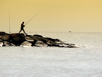 Side view of fisherman walking on rocks by sea against clear sky during sunset