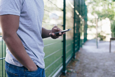 Midsection of young man standing against fence while using smart phone