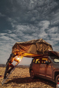 Full body side view of male camper in outerwear with burning lamp climbing on ladder in tent on roof of suv car parked in valley under cloudy sky