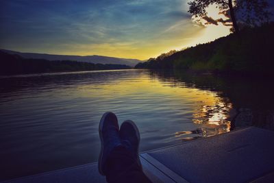 Low section of person relaxing by lake against sky during sunset