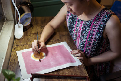From above view of crop unrecognizable female artist creating picture with paint and brush on canvas in workshop