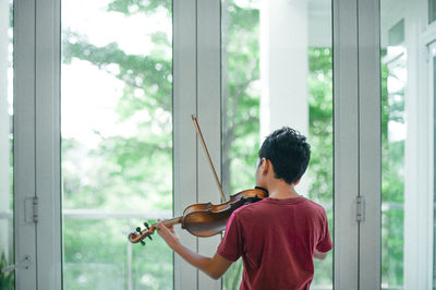 Rear view of boy playing violin by window