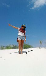 Young woman with hand raised standing at sandy beach against blue sky