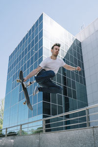 From below young guy with leg prosthesis jumping on skateboard above ground against modern building on sunny day