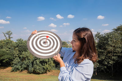 Young woman holding dart board against sky