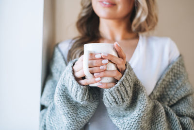 Young woman with blonde long curly hair in knitted grey sweater with cup of tea in hands at home