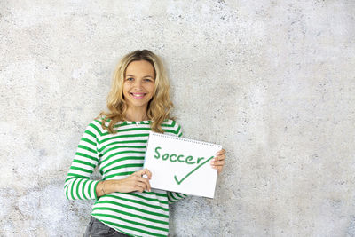 Portrait of smiling beautiful woman holding book with text while standing against wall