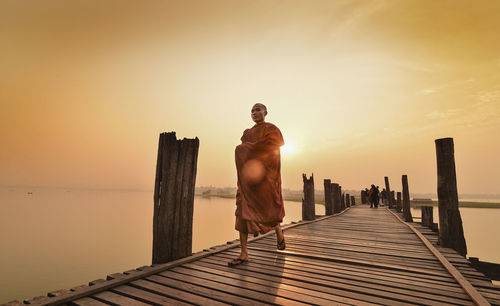 Full length of monk walking on pier over sea against sky during sunset