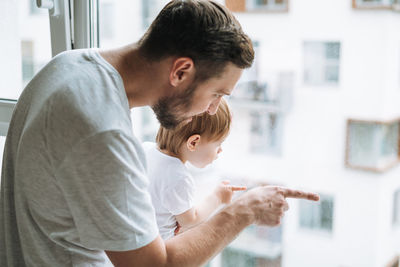 Young man father with baby girl on window sill looking at window at home