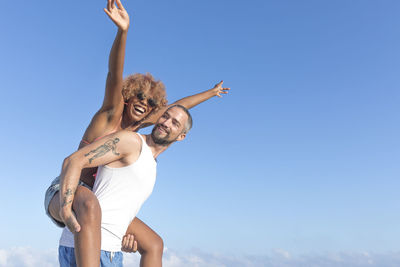 Low angle view of man piggybacking cheerful girlfriend against blue sky