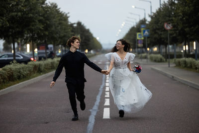 The bride in a wedding dress and the groom are running along the asphalt road with markings.