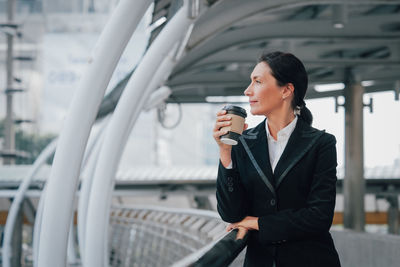 Mature woman holding coffee cup while standing by railing in city