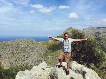 Portrait of man with arms outstretched standing on rocks against sky