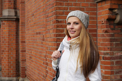 Portrait of smiling beautiful woman standing against brick wall during winter