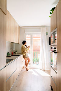 Side view of hispanic female in warm sweater with long dark hair sipping coffee and looking out window while resting in sunlit kitchen in weekend morning at home