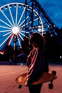 Side view of woman holding skateboard against illuminated ferris wheel