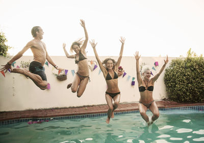 Carefree friends jumping together into swimming pool against sky during sunset