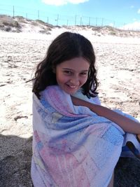 Portrait of smiling girl wrapped in towel while sitting on sand at beach
