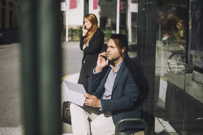 Male entrepreneur talking on smart phone while sitting with laptop at bus stop during sunny day