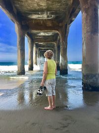 Rear view of senior woman holding sandals while standing under pier at beach