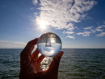 Hand holding crystal ball by sea against sky