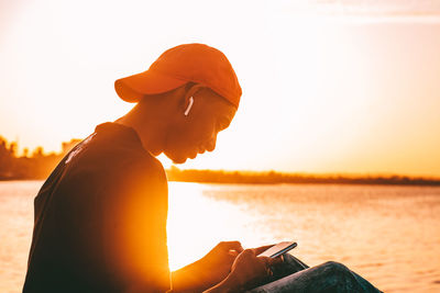 Side view of young man sitting at ocean using mobile phone against sunset