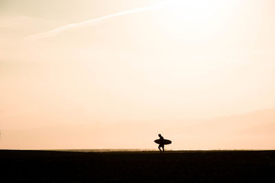 Silhouette man carrying skateboard walking on land against sky during sunset