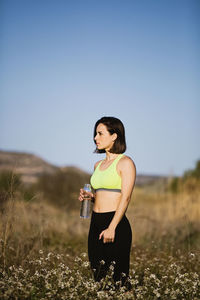 Side view of woman with water bottle standing amidst plants against clear sky