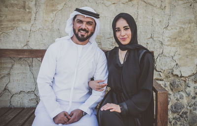 Portrait of smiling couple sitting on bench against wall