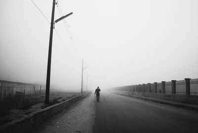 Rear view of man walking by street against sky during foggy weather