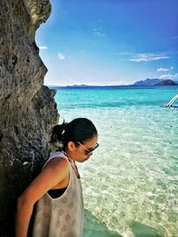 Side view of woman standing by rock at beach against blue sky