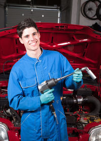 Portrait of smiling mechanic with equipment standing against car in garage