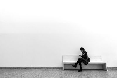 Full length of woman using mobile phone while sitting on bench against wall