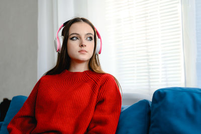 A young girl in a red knitted sweater sits on a blue sofa and listens to music in white headphones