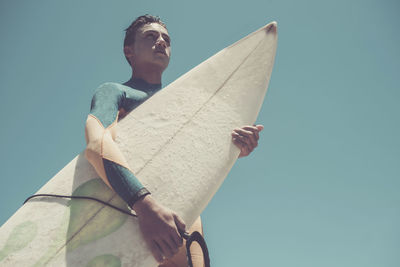 Low angle view of teenage boy holding surfboard against clear blue sky