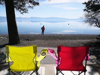 Empty chairs on walkway while man standing at lakeshore