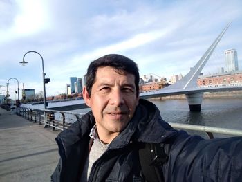Portrait of mature man sitting on bridge over river against sky