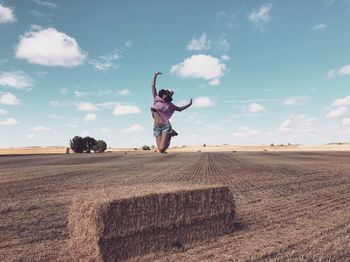 Woman with arms raised jumping over hay against sky