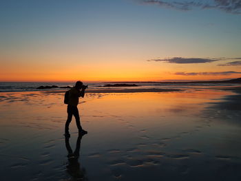 Silhouette man photographing while standing at beach against sky during sunset