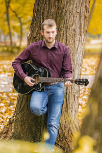 Young man playing guitar while standing by tree in park during autumn